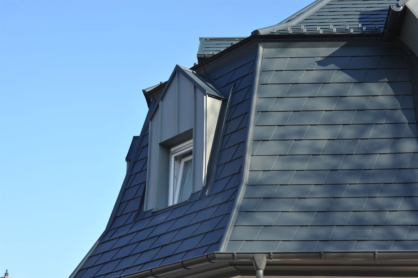 Close-up of a slate-style roof with a dormer window, gutter and downspout