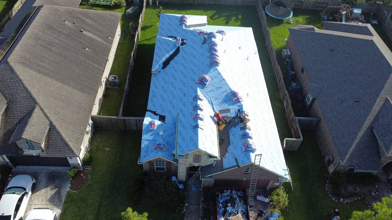 Aerial view of a home mid-replacement with blue underlayment covering the roof, shingle bundles staged along the ridge and a dumpster in the driveway